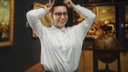 Woman wearing red glasses and white shirt smiling, hands making bunny ears near framed paintings...