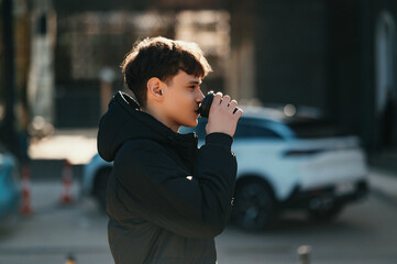 Teen White Male drinking coffee to go in Urban Street, Wearing Black Puffer Jacket And Hood. © Vulp