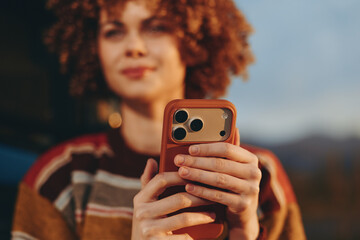 Woman with curly hair wearing a rainbow sweater smiling while holding a smartphone outdoors....