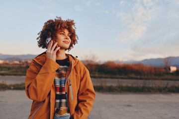 A smiling woman in a rainbow sweater and orange jacket talks on a smartphone outdoors during golden...