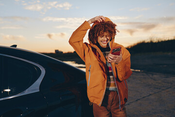 Smiling woman with curly hair wearing a rainbow sweater and orange jacket stands by a black car holding a smartphone outdoors at sunset. Lifestyle and happiness concept. © SHOTPRIME STUDIO