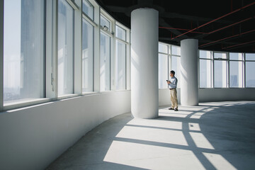 Naklejka premium Man standing by large windows in a modern unfinished office space checking his smartphone