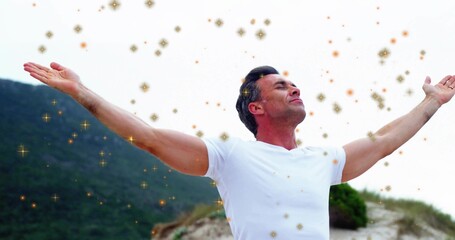 Standing mature man holding arms out on sand dune, wearing white t-shirt and golden sparkles