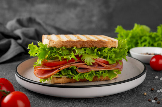 Ham and cheese sandwich with fresh lettuce and tomato on toasted bread. Close-up on a plate, juicy layers, deli style. Healthy fast food, dark background, selective focus.
