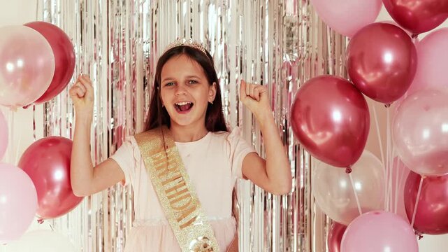 A girl is smiling and raising her arms in excitement during her birthday celebration. She is wearing a sash and surrounded by balloons and shiny decor
