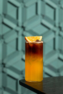 Refreshing bumble orange citrus juice and black coffee espresso drink with ice in a transparent collins highball glass on wooden table, blue tile wall background, lifestyle vertical with copy space
