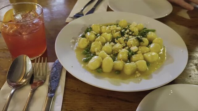 A restaurant table featuring a large plate of creamy gnocchi with cheese and sauce, paired with a refreshing cocktail.