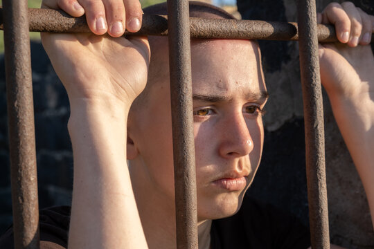 Teenage boy with shaved head stands behind prison bars holding the metal grid of a jail window and looking down with sad thoughtful expression. Concept of crime and punishment.