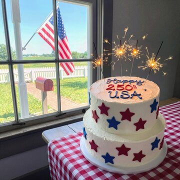 A two-tier cake adorned with red and blue stars and sparklers sits next to a window, celebrating America's 250th birthday, while an American flag gently waves outside