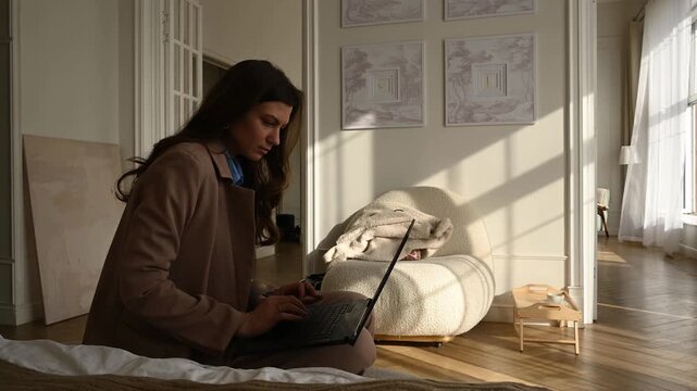 Young professional woman sitting on the floor in a sunlit apartment, concentrating on her work and typing on a laptop, embracing the freelance lifestyle and remote employment flexibility