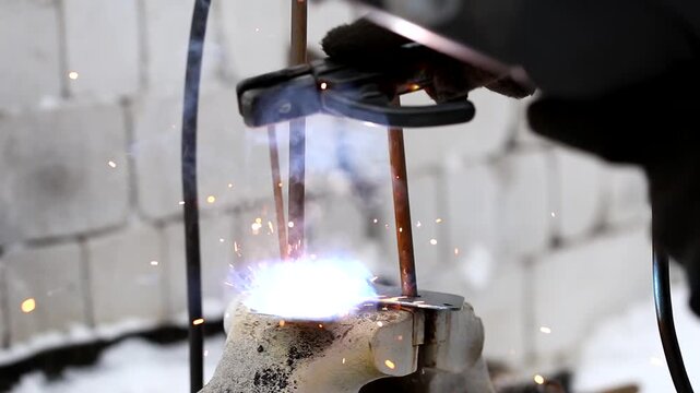 A close-up shot captures the intense glow of a welding torch as it joins two metal rods, with sparks flying and illuminating the surrounding work area.