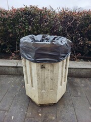 Public trash bin with black plastic bag on a city sidewalk near bushes. Urban waste disposal concept, sanitation, cleanliness and environmental management in public space