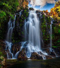 Obraz premium Large multi-tier tropical waterfall cascading over rocky cliffs into a clear pool surrounded by lush green vegetation.