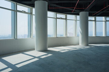Naklejka premium Spacious empty office interior with floor-to-ceiling windows, concrete columns, and dramatic light shadows