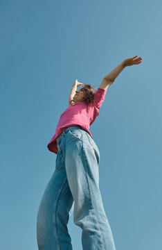 Beautiful carefree young woman with curly hair in pink t-shirt raising her hands up on blue sky background low angle shot.