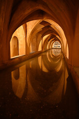 Fototapeta premium Historic underground corridor with Gothic and Moorish arches reflected in still water inside the Royal Alcazar of Seville, Spain. Atmospheric architectural perspective with symmetry, warm light 