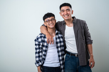 Naklejka premium Two young Asian men smiling and posing together, one in a black-and-white checkered shirt and glasses, the other in dark gray shirt and white tank top, against a plain light gray background
