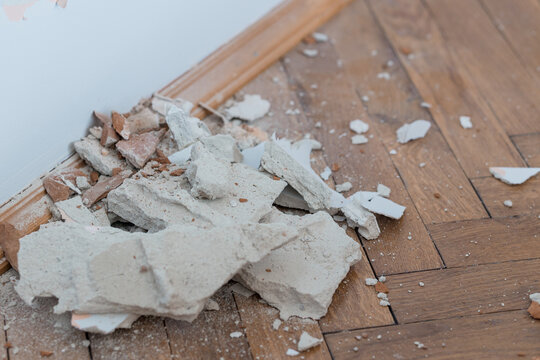 Broken plaster debris scattered on wooden parquet floor near baseboard showing renovation demolition scene with dust and rubble fragments in beige tones.