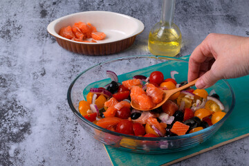 Sliced ​​ingredients for Mediterranean fish are mixed in a glass baking dish on a gray table. Raw trout fillet, olives, cherry tomatoes, red onion, herbs, capers, garlic, and olive oil. Step by step