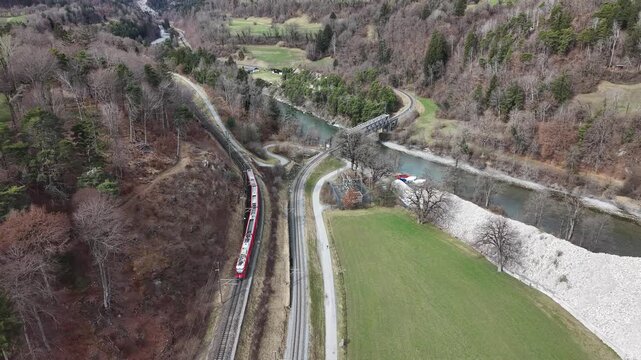 Aerial footage following a train as it curves left and ascends toward mountains, passing above a river. Captured with a drone, highlighting dynamic perspective, railway infrastructure, and natural sce