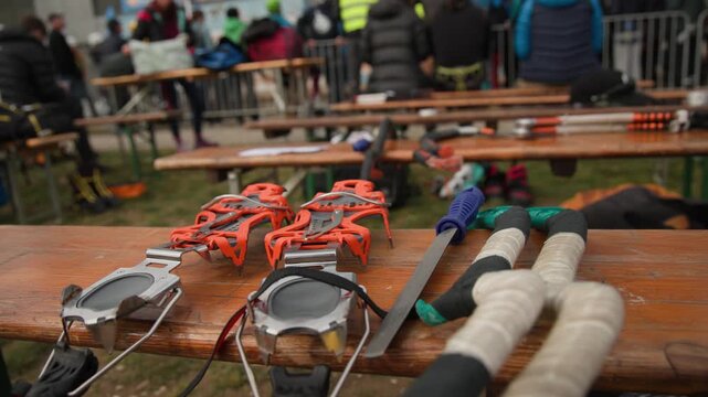Orange crampons and ice axes rest on a wooden bench, prepared for an ice climbing competition. In the background, a crowd of spectators and participants gathers, creating a focused sporting atmosphere