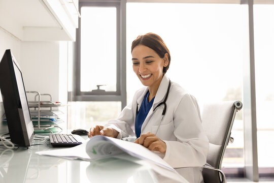 Young 35s Middle-Eastern female doctor in uniform working seated at workplace table with papers and computer, reviewing medical documents, check patient records or analyzing clinical reports on paper
