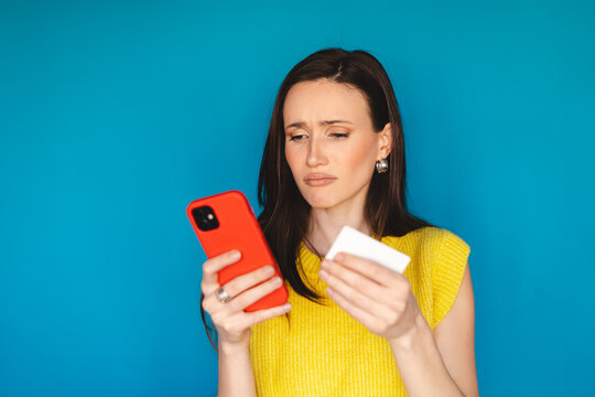 Disappointed young woman holding credit card and red smartphone, looking at screen with doubt or sadness, feeling upset by failed payment or expensive price isolated on blue background