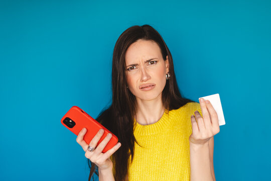  Annoyed and skeptical young woman holding credit card and red smartphone, looking at camera with grimace of disgust or disapproval isolated on blue background, bad customer experience concept
