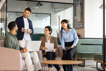 Fototapeta premium Diverse colleagues gather in office lounge with documents and laptop, discussing business project, brainstorm ideas, planning strategy during corporate teamwork Professional collaboration, partnership