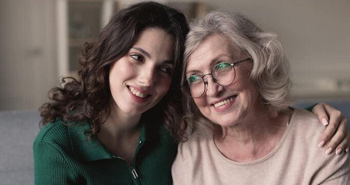 Close up portrait of young adult granddaughter hugging on shoulder senior grandmother sitting on sofa at home, spend priceless time together, expressing love, warmth. Multigenerational family bonds