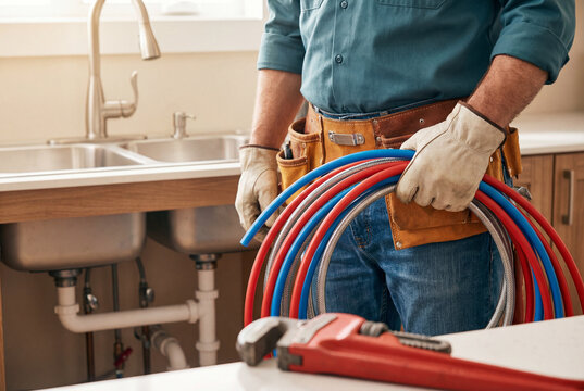 Plumber with tool belt holding colorful pipes near kitchen sink with red wrench in foreground