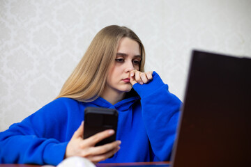 Naklejka premium Overwhelmed young woman holds smartphone while staring blankly at laptop screen. Information overload and digital distraction affect her focus. Modern anxiety and constant connectivity at home
