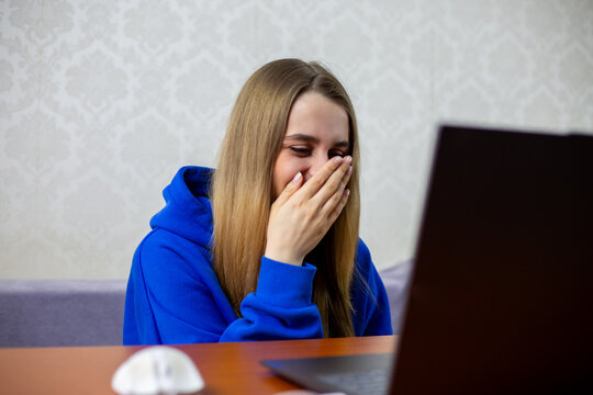 Blonde girl laughs at funny football moment on laptop, covering mouth with hand. Amusing sports blooper or unexpected play during match makes her giggle. Entertainment at home