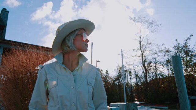Woman wearing white hat and jacket and orange glasses stands outdoors. Blonde model in stylish outfit low angle view.