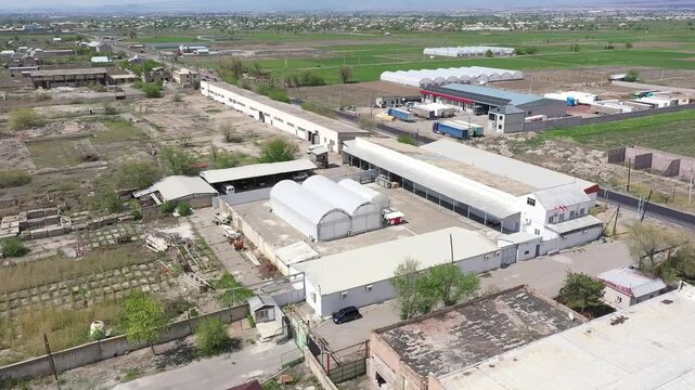 Aerial drone footage showing a large agricultural complex with multiple industrial buildings and a massive greenhouse situated in a rural landscape with green fields and a distant town.
