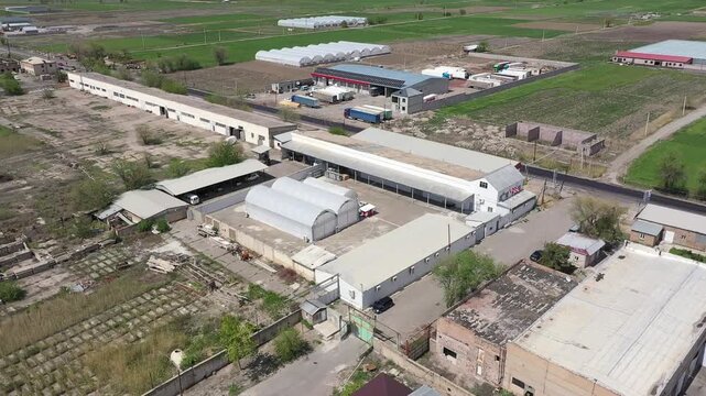 Aerial drone footage showing a large agricultural complex with multiple industrial buildings and a massive greenhouse situated in a rural landscape with green fields and a distant town.