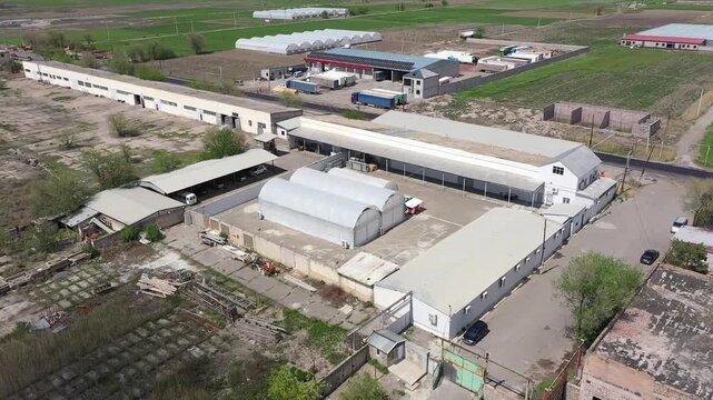 Aerial drone footage showing a large agricultural complex with multiple industrial buildings and a massive greenhouse situated in a rural landscape with green fields and a distant town.
