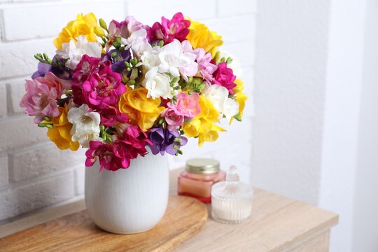 Beautiful freesia flowers in vase and candles on wooden table near white brick wall indoors, closeup. Space for text
