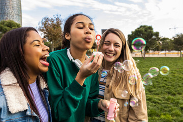 Fototapeta premium Three young diverse women friends enjoying a joyful day outdoors in a park, blowing soap bubbles and laughing together, sharing a moment of carefree connection and happiness