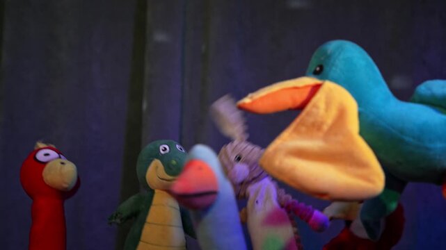 Three colorful bird puppets or toy figurines dance under blue stage lighting next to a glowing yellow lampshade, creating a whimsical theatrical scene.