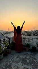 Fototapeta premium Woman in red dress celebrating victory with raised arms on urban rooftop terrace at golden sunset over city skyline with communication towers and buildings.