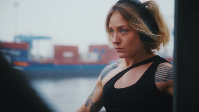 Tattooed Athletic Woman Doing Dumbbell Front Raise with Headphones at Industrial Harbor Waterfront