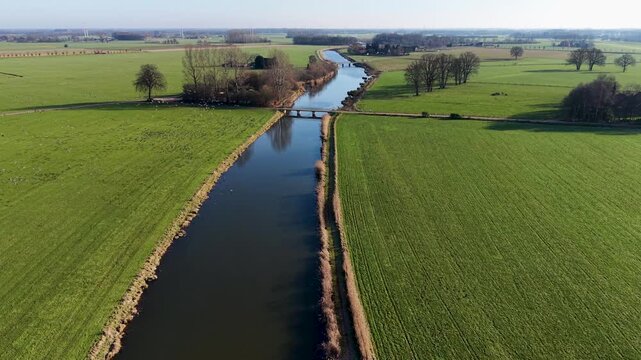 Aerial view of a rural landscape with a canal curving through green fields, two small road bridges crossing it, and a nearby house with outbuildings set among open farmland.
