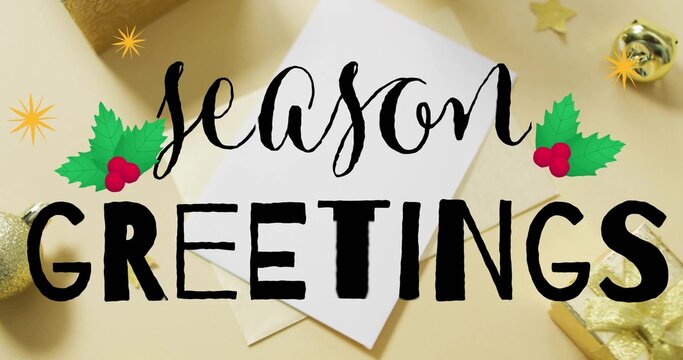 Showing white card and envelope with black season GREETINGS text on beige tabletop, gold bauble