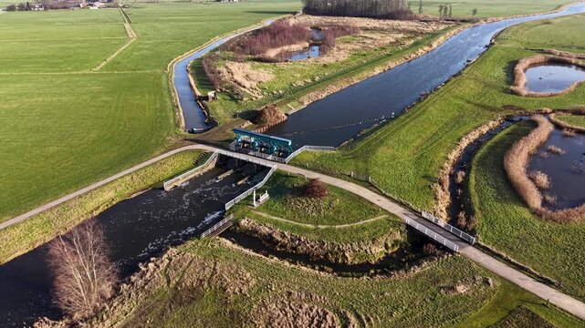 Aerial drone view of a straight Dutch canal and parallel cycling path cutting through lush green polder landscapes, featuring a blue water control gate and a traditional farmhouse in spring.