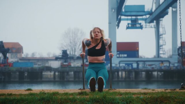 Tattooed Athletic Woman Doing Tricep Dips on Parallel Bars at Industrial Harbor Waterfront