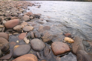 Smooth river stones on a tranquil bank where clear water gently laps against the shore, creating a peaceful natural scene in the serene outdoors, reflecting nature's calm beauty