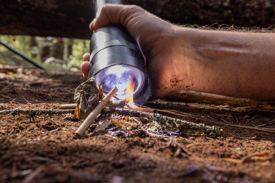 Powerful flashlight starts a fire on kindling. Fingers grip flashlight, steady, focused. Fire crackles low beneath forest floor. Dirt, twigs, and leaves surround glowing flame