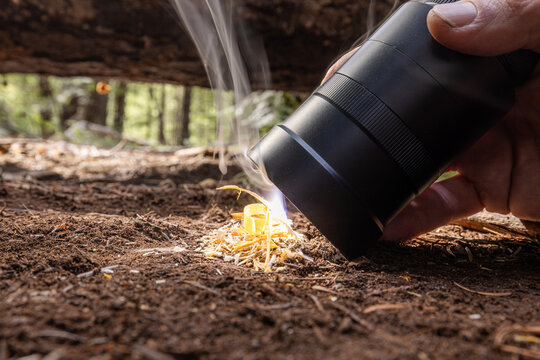 Powerful flashlight starts a fire on kindling. Fingers grip flashlight, steady, focused. Fire crackles low beneath forest floor. Dirt, twigs, and leaves surround glowing flame