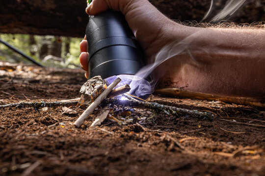 Powerful flashlight starts a fire on kindling. Fingers grip flashlight, steady, focused. Fire crackles low beneath forest floor. Dirt, twigs, and leaves surround glowing flame